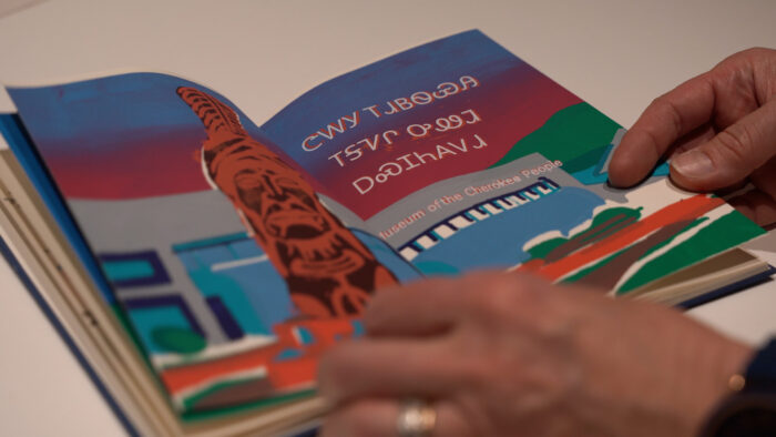 Someone's hands holding a book in the Cherokee language and a sentence in English that reads, "Musuem of the Cherokee People."