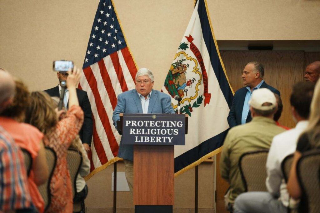 A man in a suit and collared shirt stands at a podium before an audience of people sitting. He stands in front of the American and West Virginia flags and speaks into a microphone.