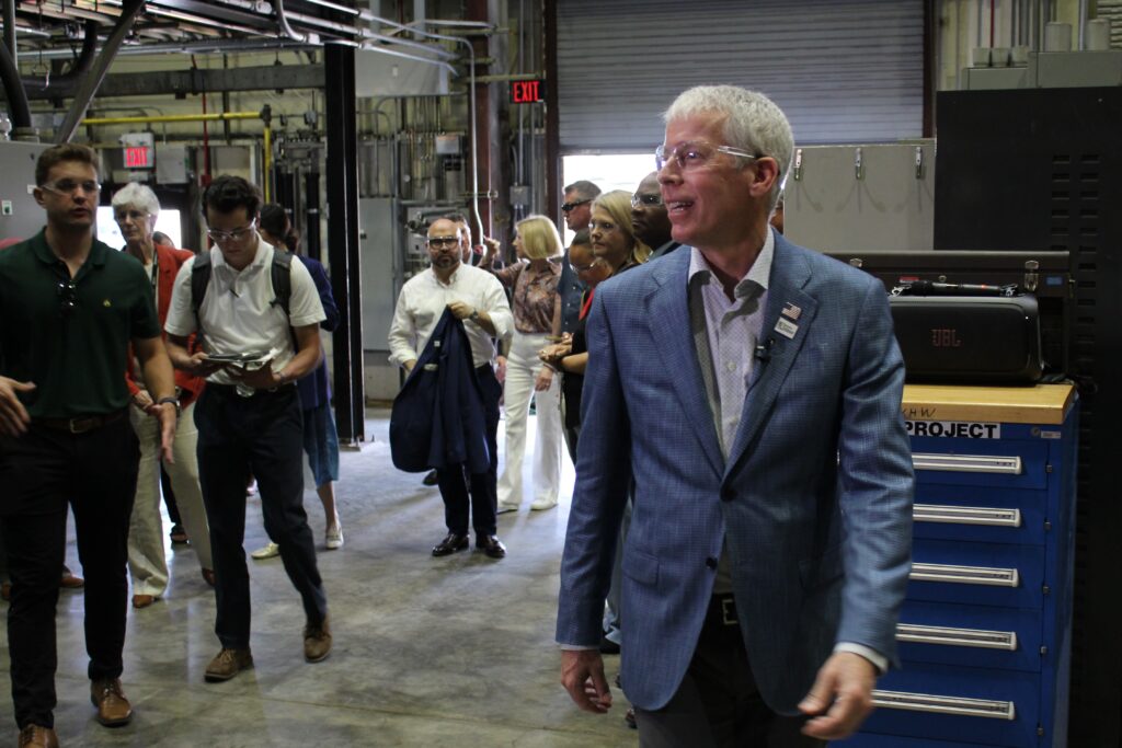 A man with short white hair wearing safety glasses, a light blue suit jacket over an off-white shirt and charcoal grey pants walks towards the lens while facing frame left. Behind him several people stand dressed in business casual and formal clothes. All are in a large warehouse in front of a bay door rolled up and letting in daylight.