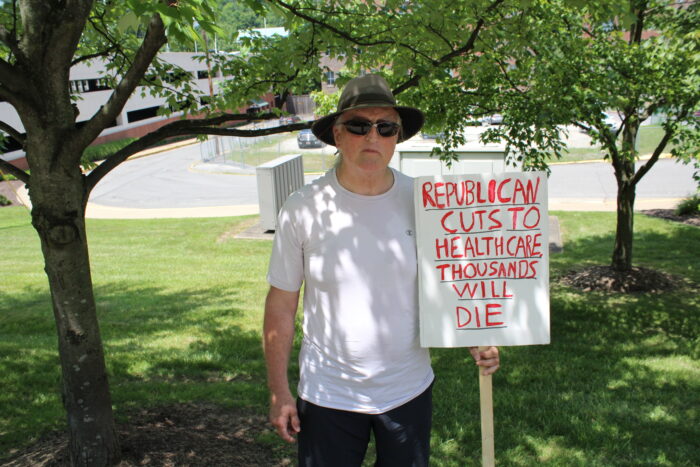 A man wearing a bucket hat and sunglasses and a white shirt holds a sign that reads "Republican Cuts to Healthcare Thousands Will Die"