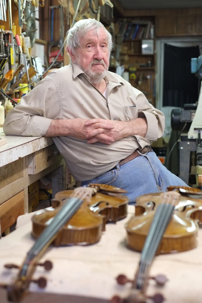 A white man leaning on a counter in his workspace in front of two fiddles.