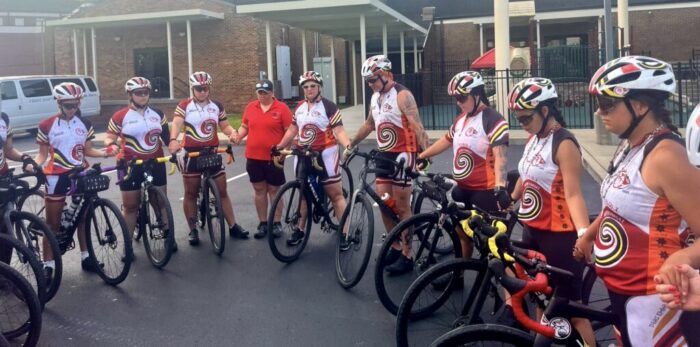 Over 10 cyclists holding hands in a circle outside a building. They are wearing uniforms with white, red, yellow and orange accents.