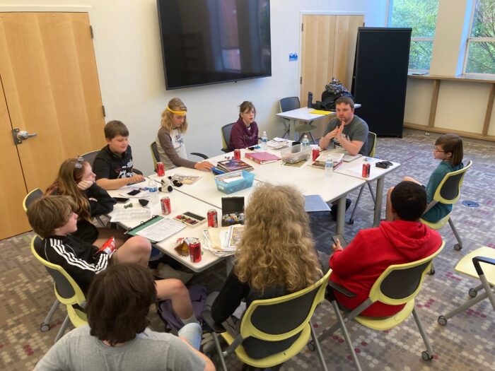 Nine kids sitting in a semi-circle around a table. They are listening to an adult man explain something. The table is covered in game pieces for Dungeons and Dragons.