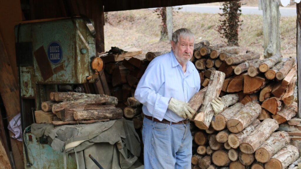 A man holding a log of wood in front of a stack of logs.