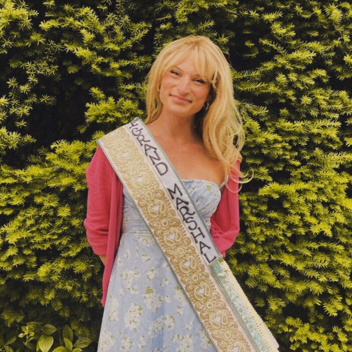 A transgender woman with long blond hair wearing a dress and pink sweater poses for a photo smiling.