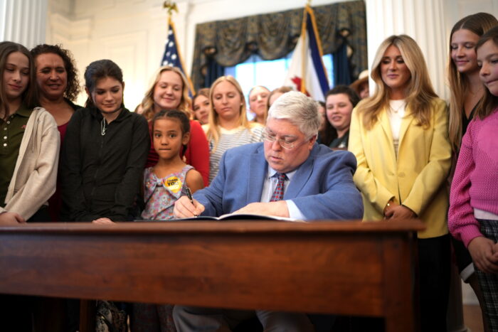 A man with white hair signs a document at a desk. Around him stand young women.