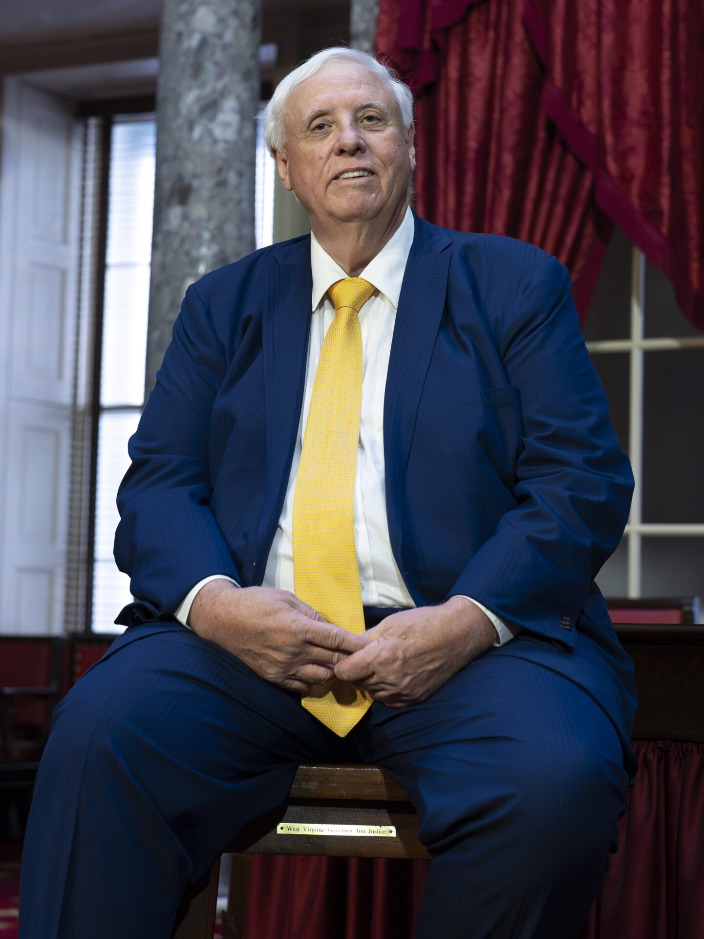 A large. white-haired man in a blue suit with a gold tie sits smiling on a desk in front of two tall, ornate columns and a window with red drapes.