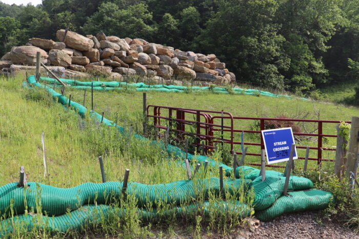 Temporary barriers to divert stormwater runoff curt across a grassy field with a large pile of rocks behind them. A blue sign marks where a gas pipeline crosses under a gravel road.