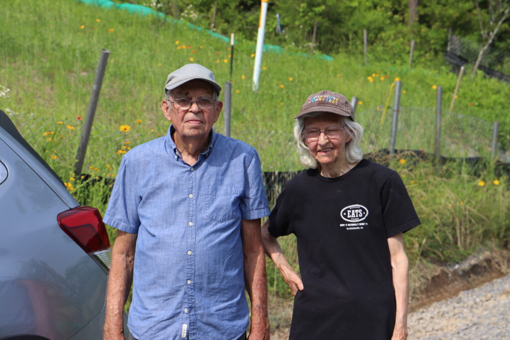 An older man and woman, both wearing hats, stand together on a gravel road on a warm summer day, with a grassy slope behind them.