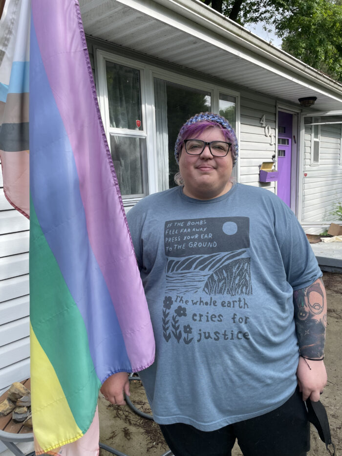 A transgender person stands on the porch of a house holding up a Pride flag. They smile.