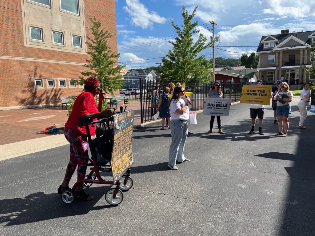 A woman wearing a bright red jacket and hat holds a sign in protest, surrounded by other people holding signs in protest on a warm summer afternoon.