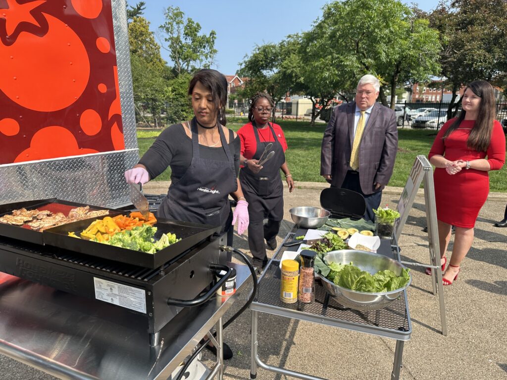 A woman cooks fresh vegetables on a grill as another woman holds a spatula while a man in a business suit and a woman in a red dress look on.