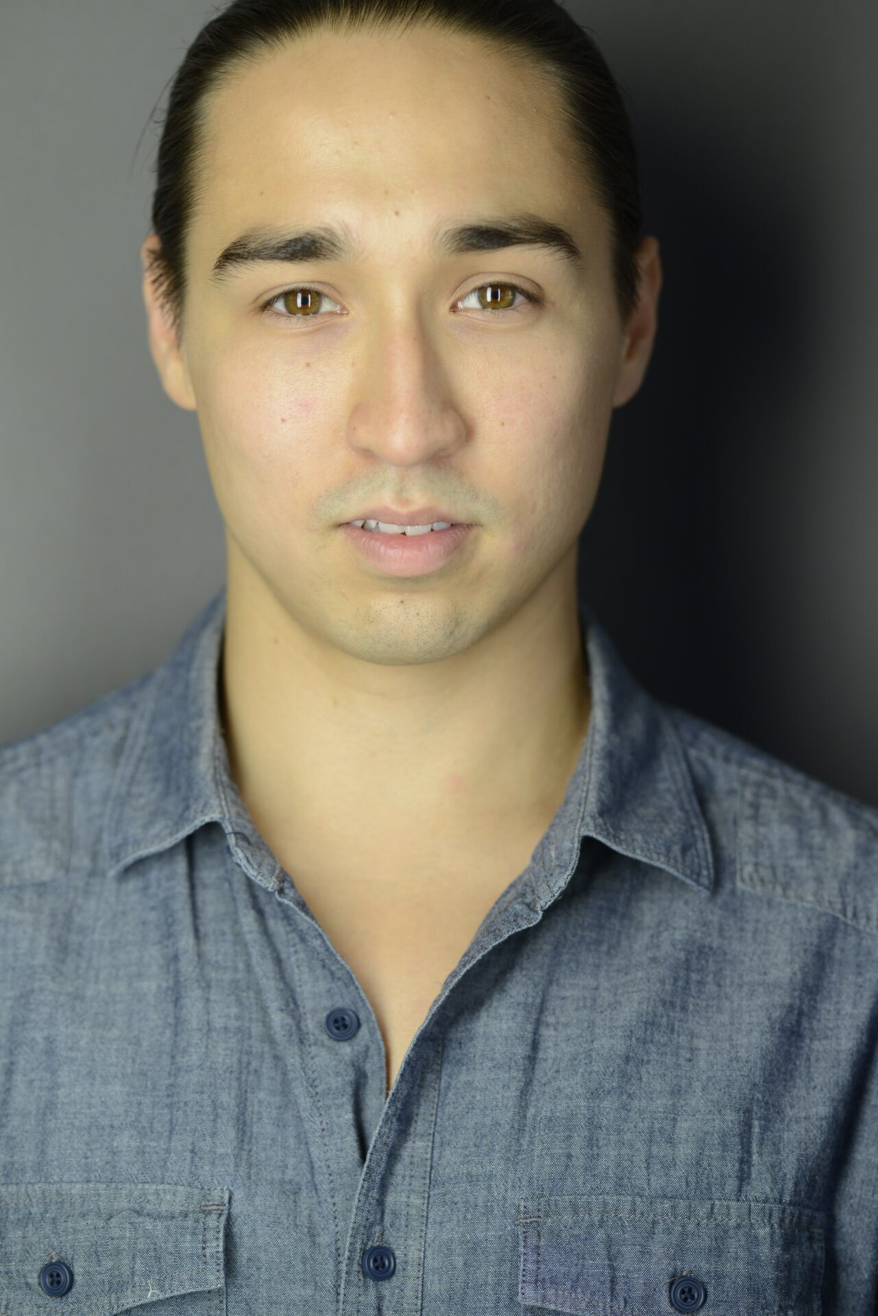 A man with a button-up shirt stares at the camera, standing before a solid-color backdrop. It is a portrait-style photo.