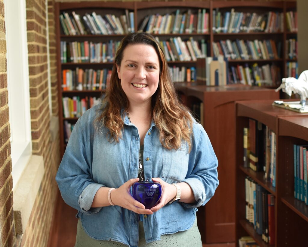 A woman is smiling. She has long brown hair and wearing a jean jacket holds the Blenko Above and Beyond blue glass apple award.