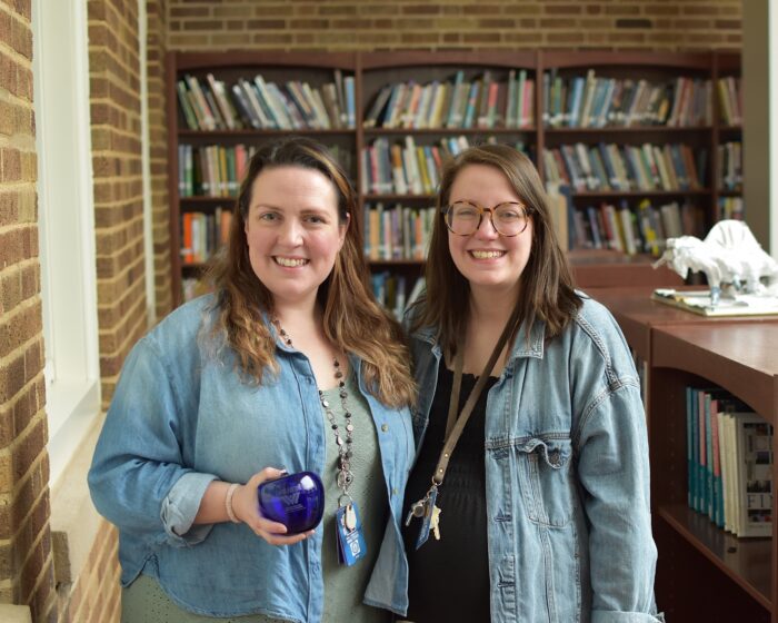 Two women are standing next to each other with books in the background. The woman on the left is holding the Above and Beyond award which is a blue glass apple from Blenko Glass.