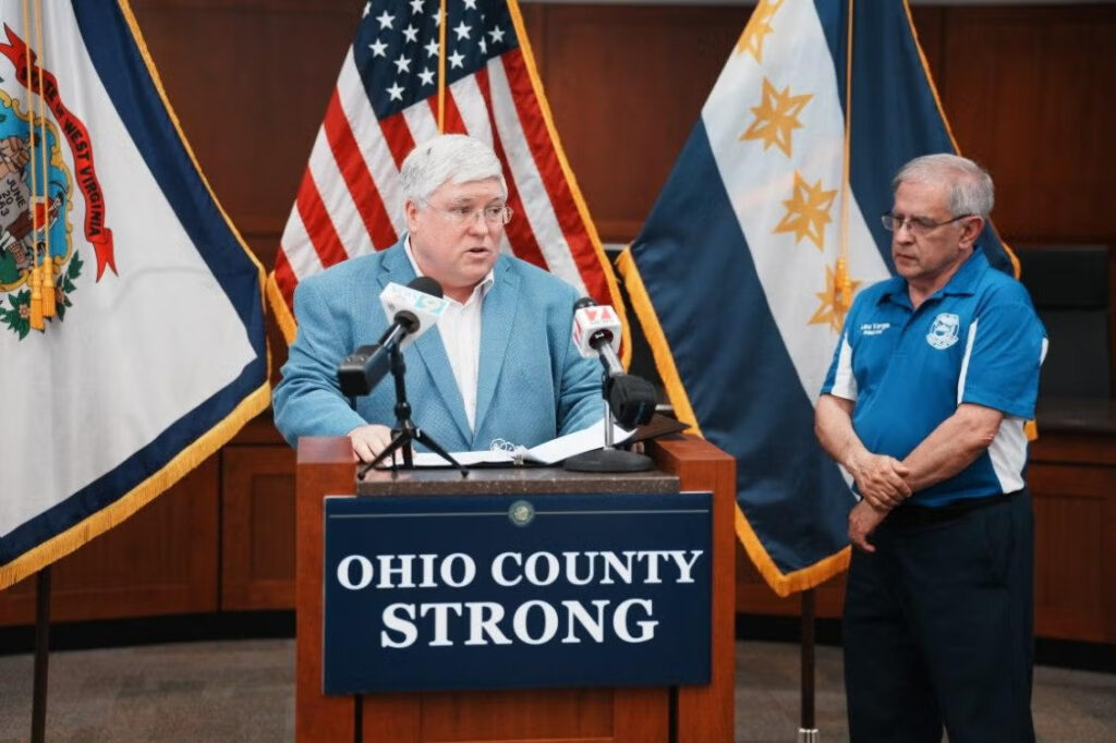 A man in a suit stands at a podium and speaks into a pair of microphones. He has a binder out with sheets of paper in front of him. On the podium, a sign reads "Ohio County Strong." Beside the podium stands another man in a collared shirt. In the background, the West Virginia and American flags are visible.