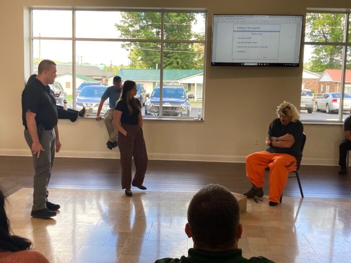 Two people standing and one person sitting in a chair in a room. Other people are watching the interaction.