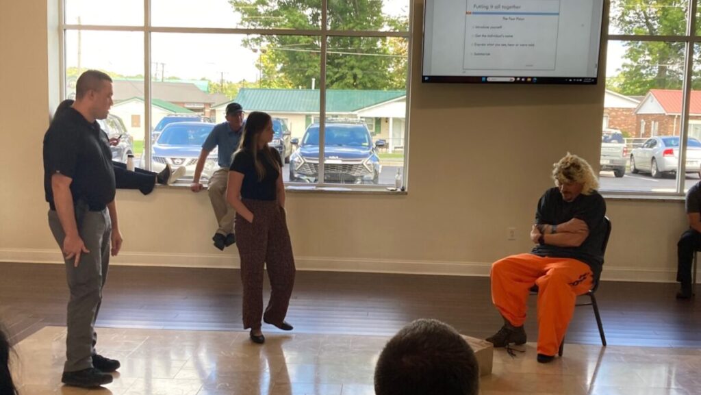 Two people standing and one person sitting in a chair in a room. Other people are watching the interaction.