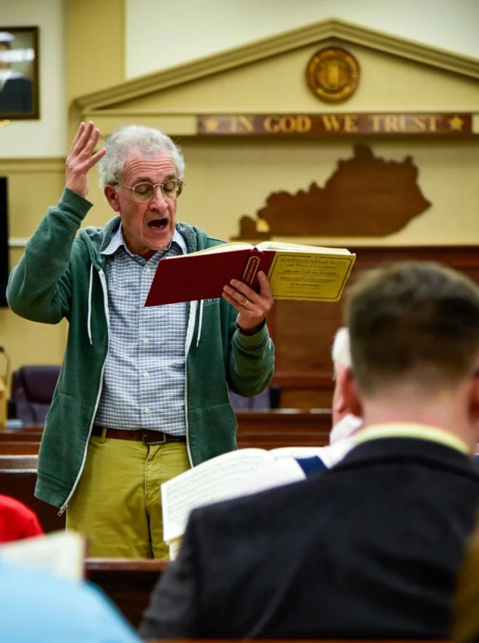 A person standing holding a book with one hand in the air. The outline of the state of Kentucky hangs on a wall behind them.