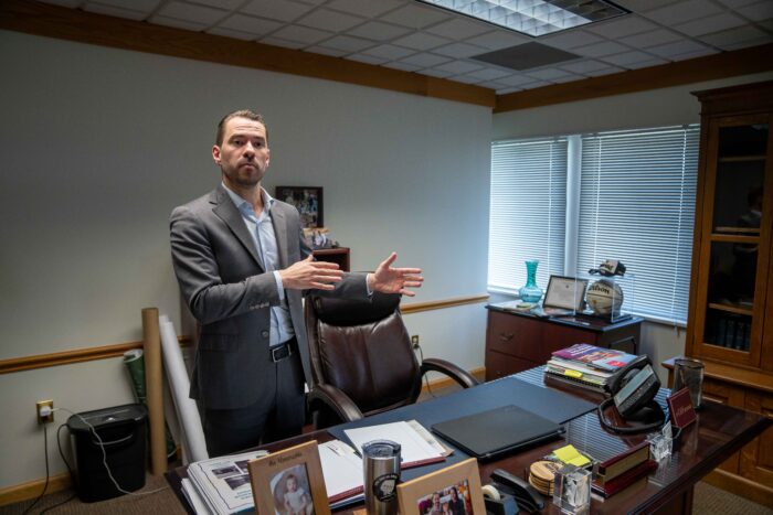 A man in a suit over a light blue button down shirt with no tie stands behind a desk in an office. He gestures with both hands to the right of frame