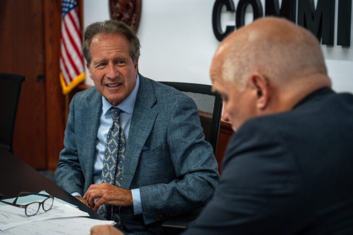 Two suited men speak behind a desk