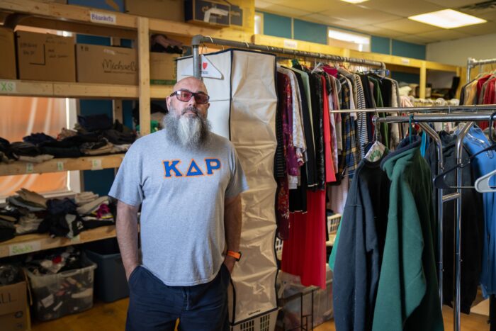 Man in a t-shirt with Greek letters on it stands around clothing on racks and shelves.