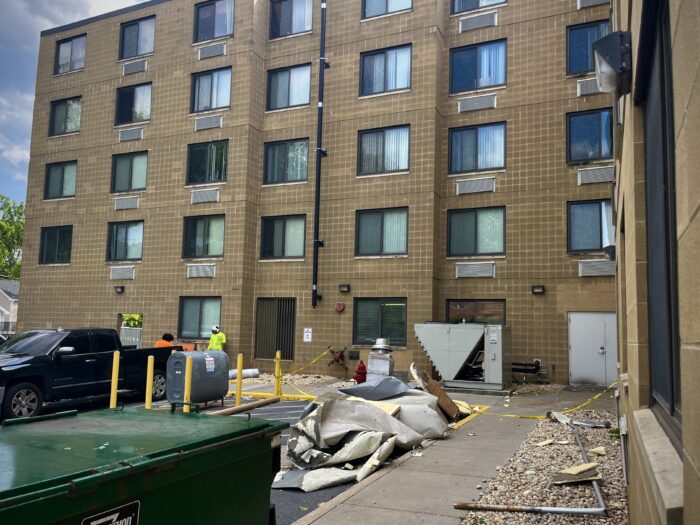 A modest amount of debris lines the sidewalk surrounding an apartment complex. Some of it is sealed off behind yellow "caution" tape. Some people in neon shirts stand nearby, likely part of the repairs team.
