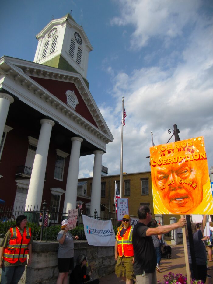 People holding picket signs, one depicting the face of Donald Trump, stand outside the Charles Town courthouse, a tall, red building with white columns.