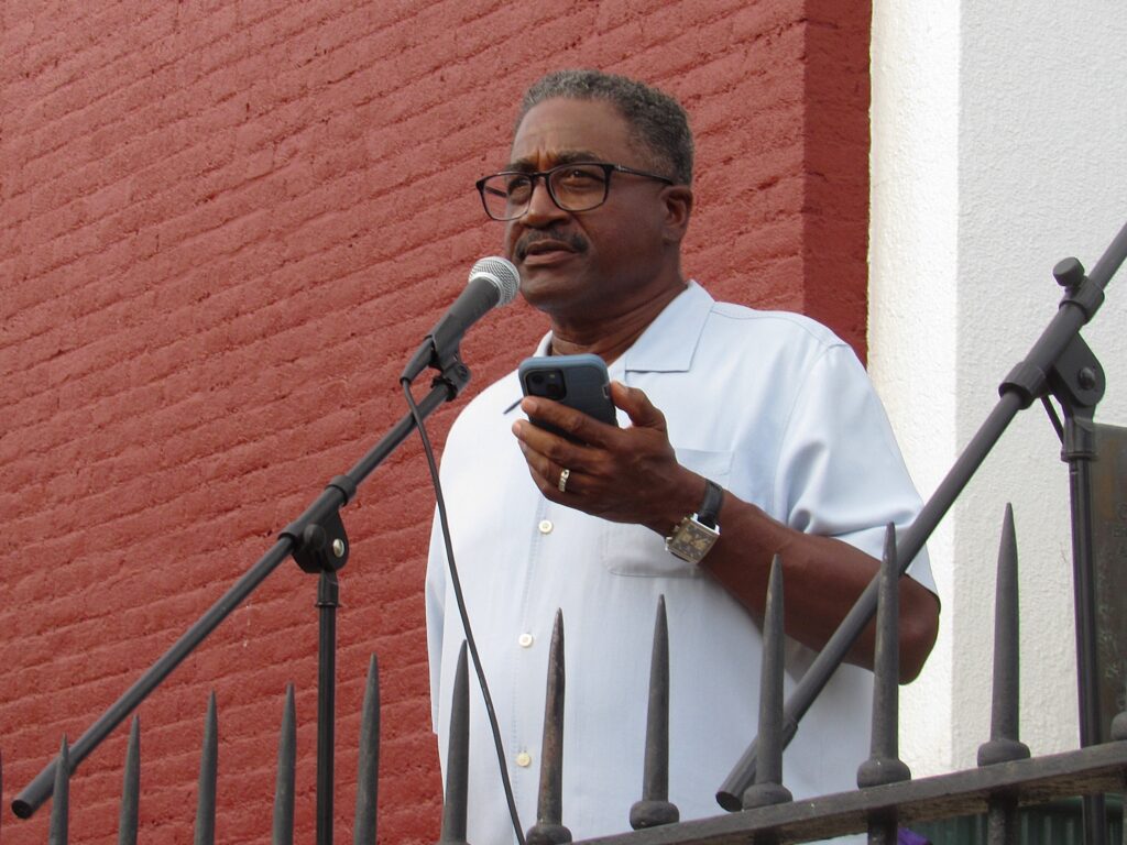 A man in glasses and a collared shirt stands and speaks into a microphone, looking forward. In his left hand he holds a cell phone.