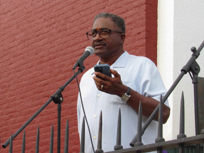 A man in glasses and a collared shirt stands and speaks into a microphone, looking forward. In his left hand he holds a cell phone.