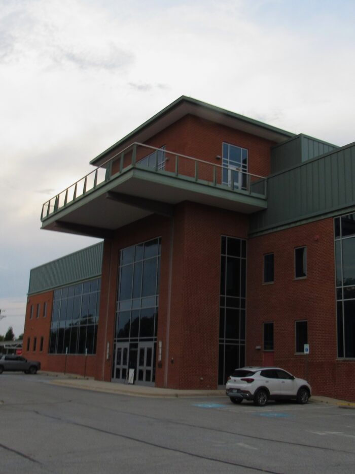 A red brick building has large windows, green panel roofing and a parking lot out front.
