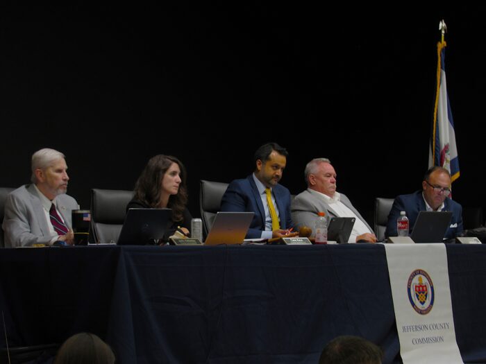 Five people in formal attire sit at a desk with microphones and a banner that reads "Jefferson County Commission."