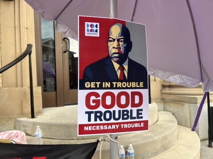 An illustration of John Lewis in a suit and tie is located on a poster that reads "Get In Trouble. Good Trouble. Necessary Trouble." It is hanging on an umbrella outside a building.