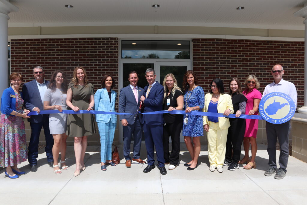People line up outside a brick building to cut a ribbon. They are dressed in formal attire and smiling toward the camera.