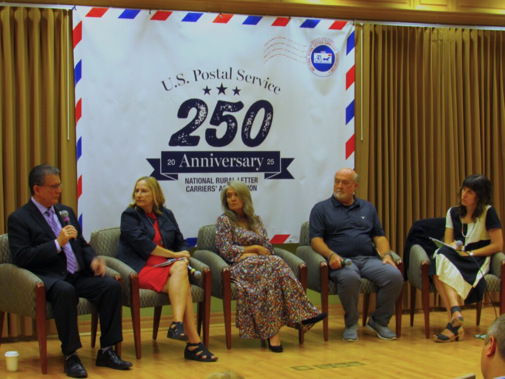 A group of people in semi formal attire sit on a stage holding microphones. Behind them a sign reads "U.S. Postal Service 250 Anniversary."