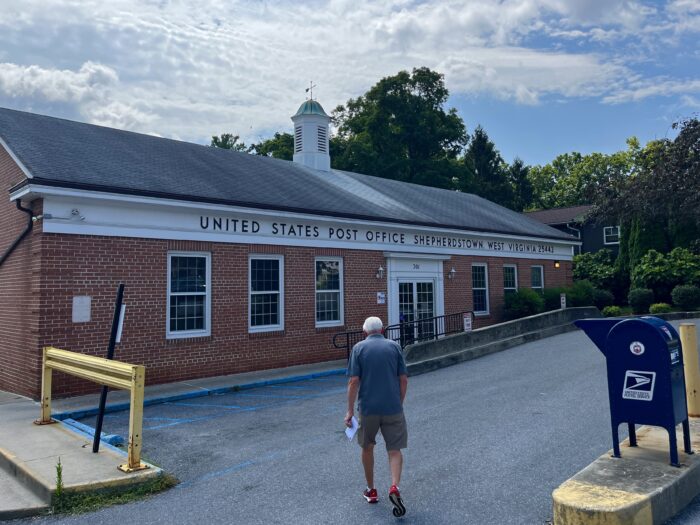 A man holding an envelope walks toward a long brick building labeled "United States Post Office Shepherdstown, West Virginia." Outside, a blue mailbox is visible.
