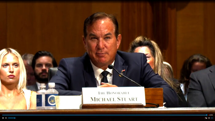 Man sits at a desk in a dark suit with a name placard in front of him