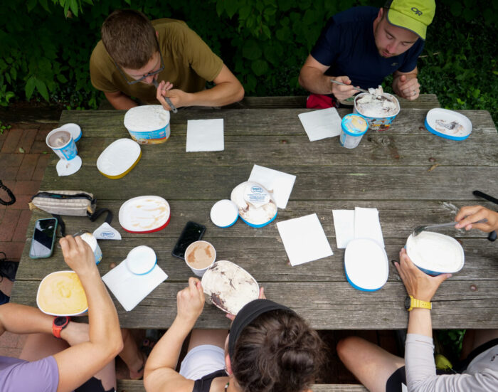 A view from above of five people sitting around a picnic table eating from pints and half gallons of ice cream.