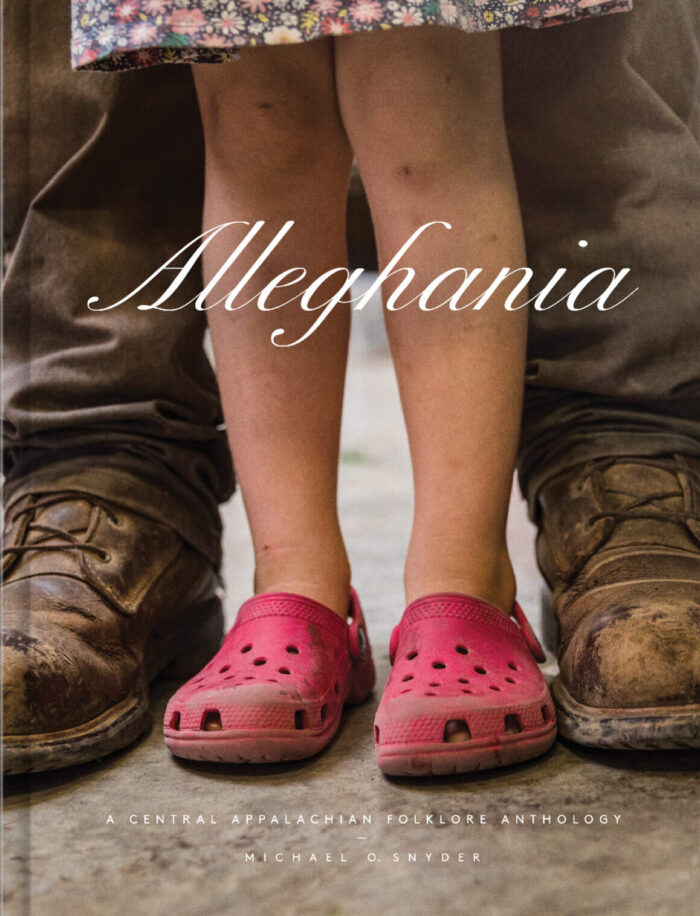 A book cover with a photograph of an adult's legs wearing brown pants and boots. A child is standing in front of them, but only their legs are visible. They're wearing pink crocs. The title on the cover reads, "Alleghania: A Central Appalachian Folklore Anthology." The author is Michael Snyder.