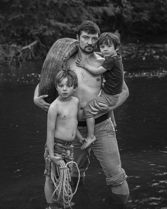 A black and white photo of a man standing shirtless in a creek holding a child and a tire around his shoulder. Another child is standing in front of him with a rope.