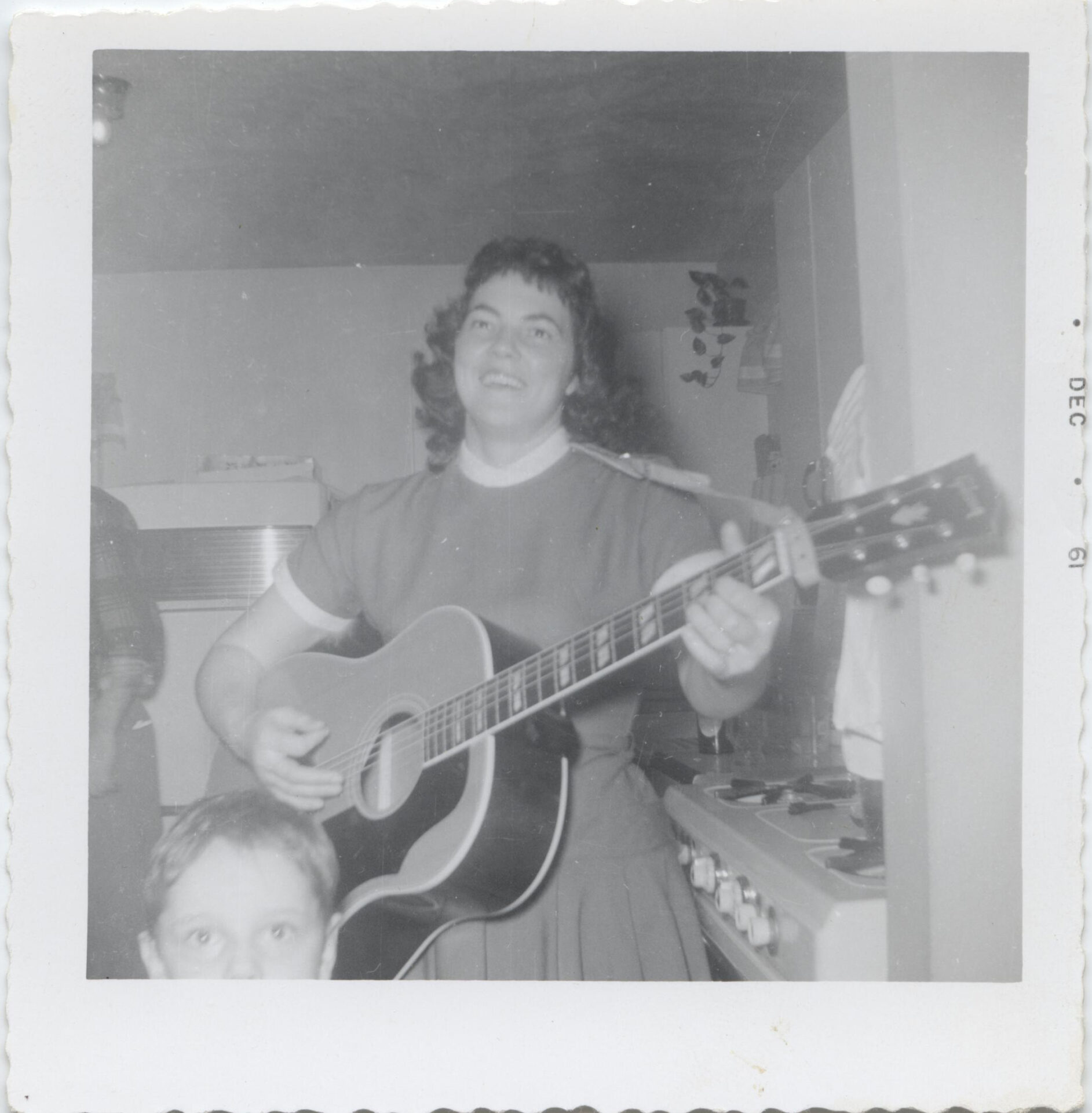A black-and-white polaroid photo of a woman holding a guitar. There is a child in the corner with only the upper half of their head visible. 