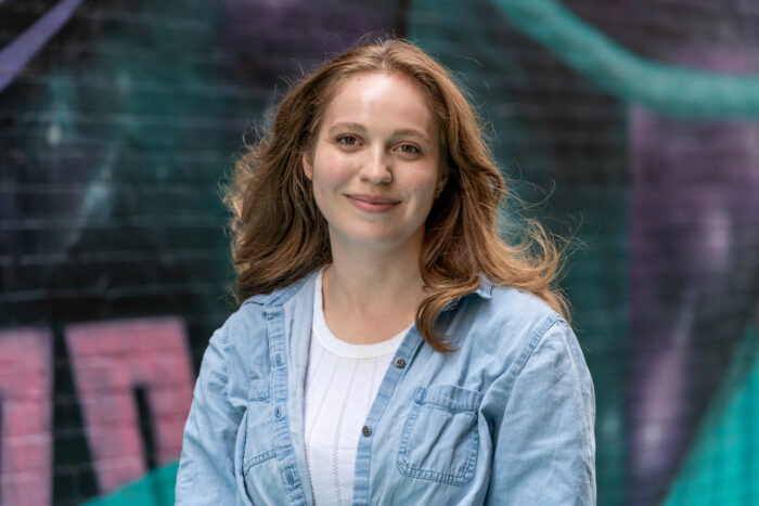 A young white woman with red hair smiles for the camera. She stands in front of a brick wall with colorful graffiti. She wears a light blue jean jacket over top a white shirt.