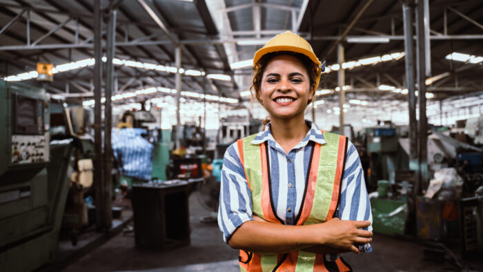 A young woman in a yellow hard hat and safety vest stands in front of industrial workers.