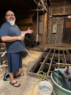 Man in shorts stands by a hole in the floor caused by flooding. 