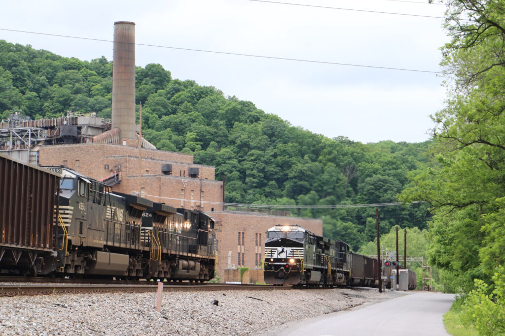 Two trains with black locomotives pass each other in front of a large brick building with a large smokestack on an overcast day.