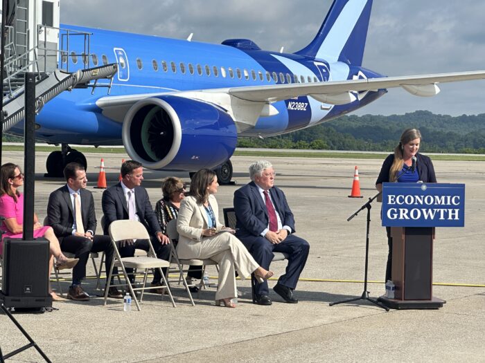 A woman in a professional suit speaks from an outdoor podium with a large airport in the background and two rows of professionally attired men and women seated to the left of the photo.