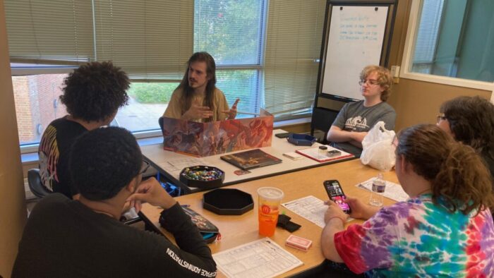 Five teenagers sitting around a table listening to an adult explain how to play Dungeons & Dragons.