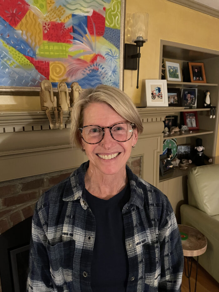 A middle age white woman with blond hair and wide-rimmed glasses. She has a nose piercing. She smiles for the camera and wears a checker patterned button up over a black shirt. Behind her is a brick fireplace and a colorful painting.