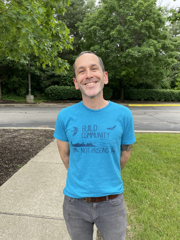 A middle age white man smiles for the camera. He is outside standing on a sidewalk. Behind him is a parking lot and green trees. He has very short trimmed hair, has his hands behind his back and wears a blue t-shirt that reads, "Build Community Not Prisons."