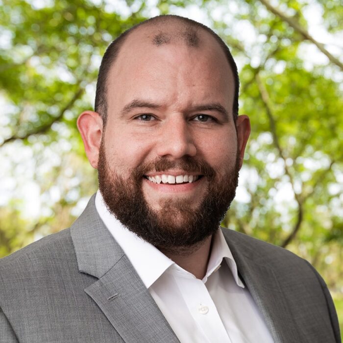 A professional headshot of a middle age white man with a brown beard and mustache. He wears a gray blazer over top a white button up shirt.